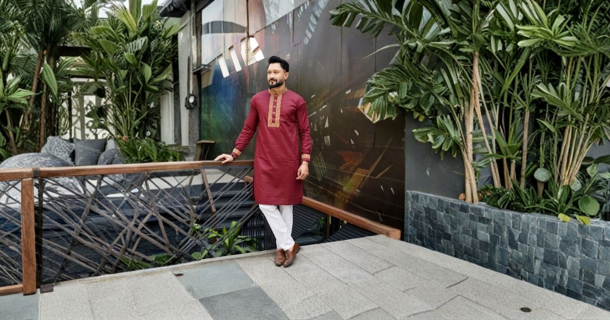 Rajvansh - Best Nawabi Kurta Designs for Weddings and Celebrations - Rajvansh Man wearing maroon kurta and white pants stands by a wooden railing in a lush indoor garden space.