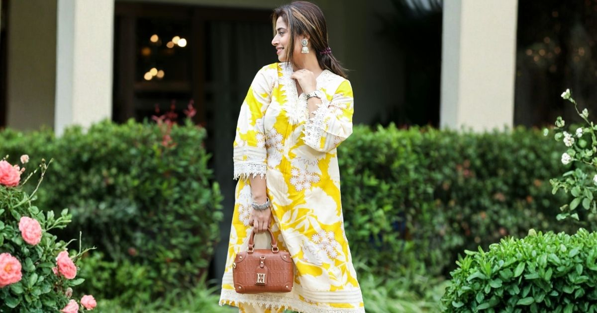 Woman in a yellow and white floral tunic dress stands in a garden, holding a brown handbag with a quilted pattern.