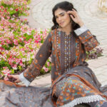 Woman in a grey and orange traditional dress sits on a brick path beside a bed of pink and white flowers, posing gracefully with one hand touching her hair.