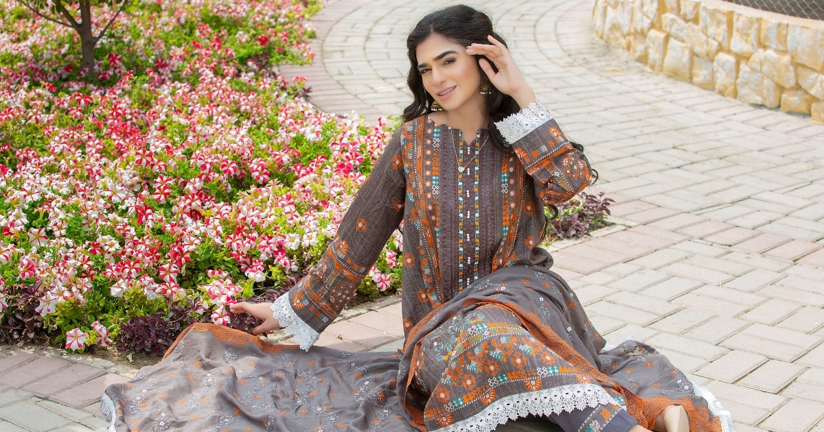 Rajvansh - Why Unstitched Suit Fabric for Women Remains Popular - Rajvansh Woman in a grey and orange traditional dress sits on a brick path beside a bed of pink and white flowers, posing gracefully with one hand touching her hair.