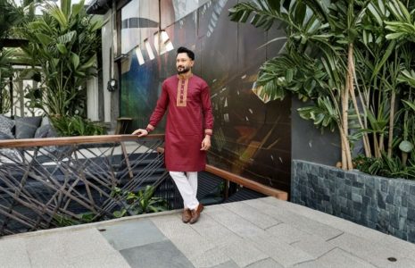 Man wearing maroon kurta and white pants stands by a wooden railing in a lush indoor garden space.