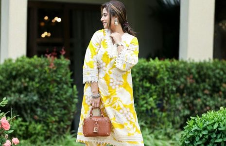 Woman in a yellow and white floral tunic dress stands in a garden, holding a brown handbag with a quilted pattern.