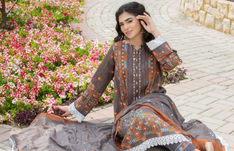 Woman in a grey and orange traditional dress sits on a brick path beside a bed of pink and white flowers, posing gracefully with one hand touching her hair.